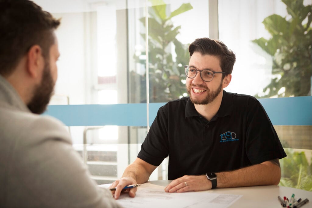 A smiling man wearing glasses and a black polo shirt with a "Be Shaw Drafting" logo sits at a table, discussing architectural blueprints with a client.