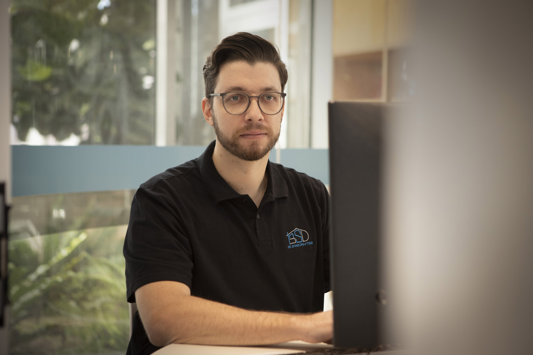 A man wearing glasses and a black polo shirt with a "Be Shaw Drafting" logo sits at his desk, looking slightly past the side of a computer monitor positioned in the foreground.